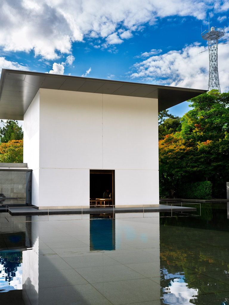 A stark white cube building beside a still reflecting pool, with a single dark opening at ground level.